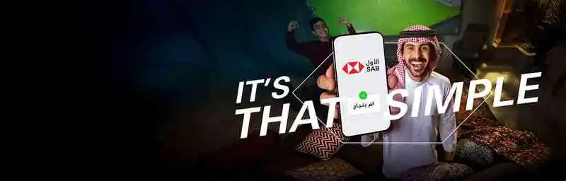 Saudi guy in a living room holding his phone towards the camera and behind him his son and a TV screen showing a soccer match