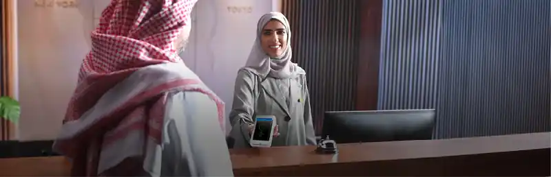 The image shows a receptionist smiling while showing a mobile phone to a man wearing traditional attire.